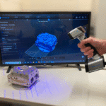 Handheld 3D scanner capturing part on desk, scanned point cloud visible on screen prior to meshing.