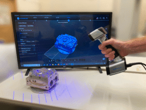 Handheld 3D scanner capturing part on desk, scanned point cloud visible on screen prior to meshing.