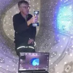 Man in enclosed space holding 3D scanner with laptop in foreground showing scan data.