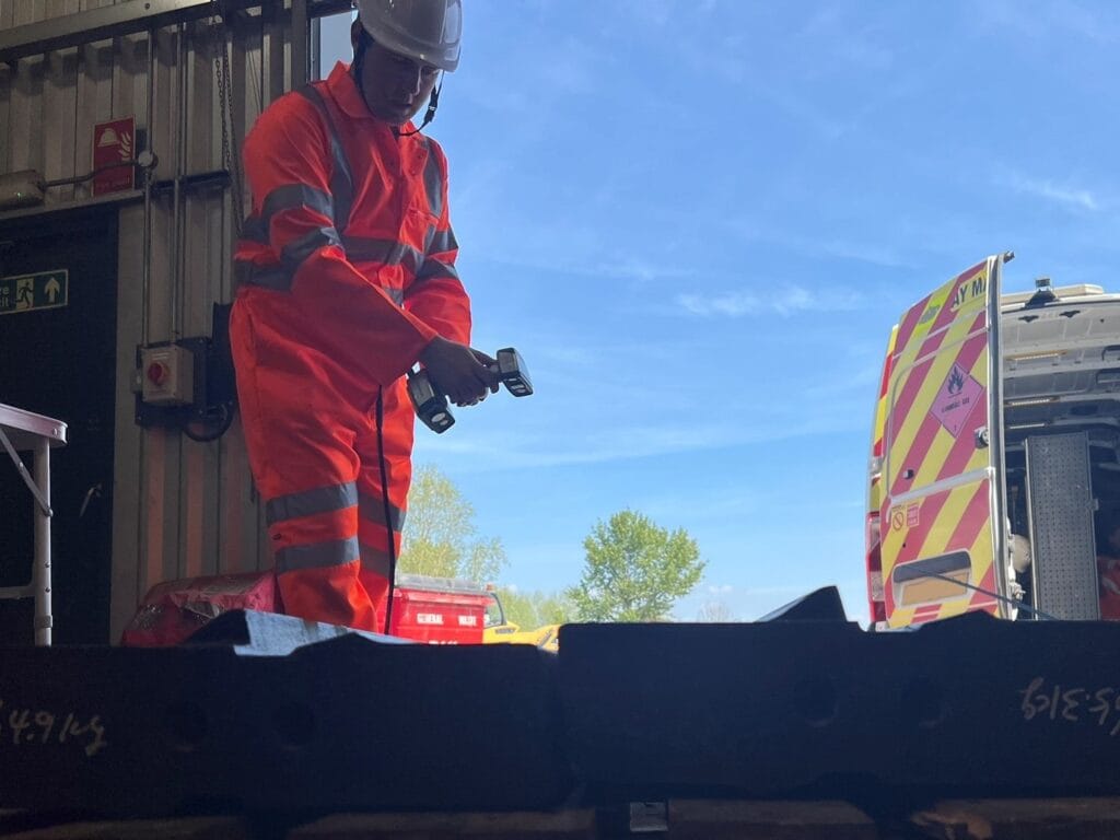 Engineer in bright orange PPE and helmet using a handheld 3D scanner on a part at a customer site.