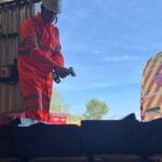 Engineer in bright orange PPE and helmet using a handheld 3D scanner on a part at a customer site.