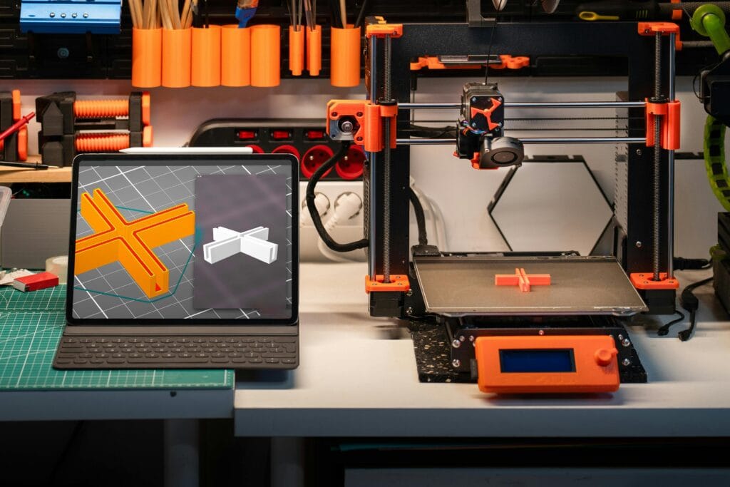 Black and orange desktop 3D printer beside tablet showing 3D model.