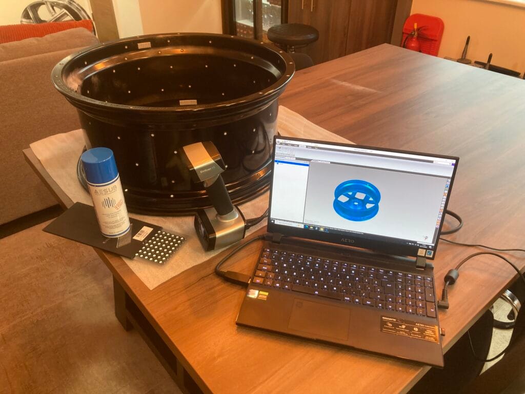 Photo of black alloy wheel, handheld 3D scanner, spray, and laptop showing 3D scanned mesh.