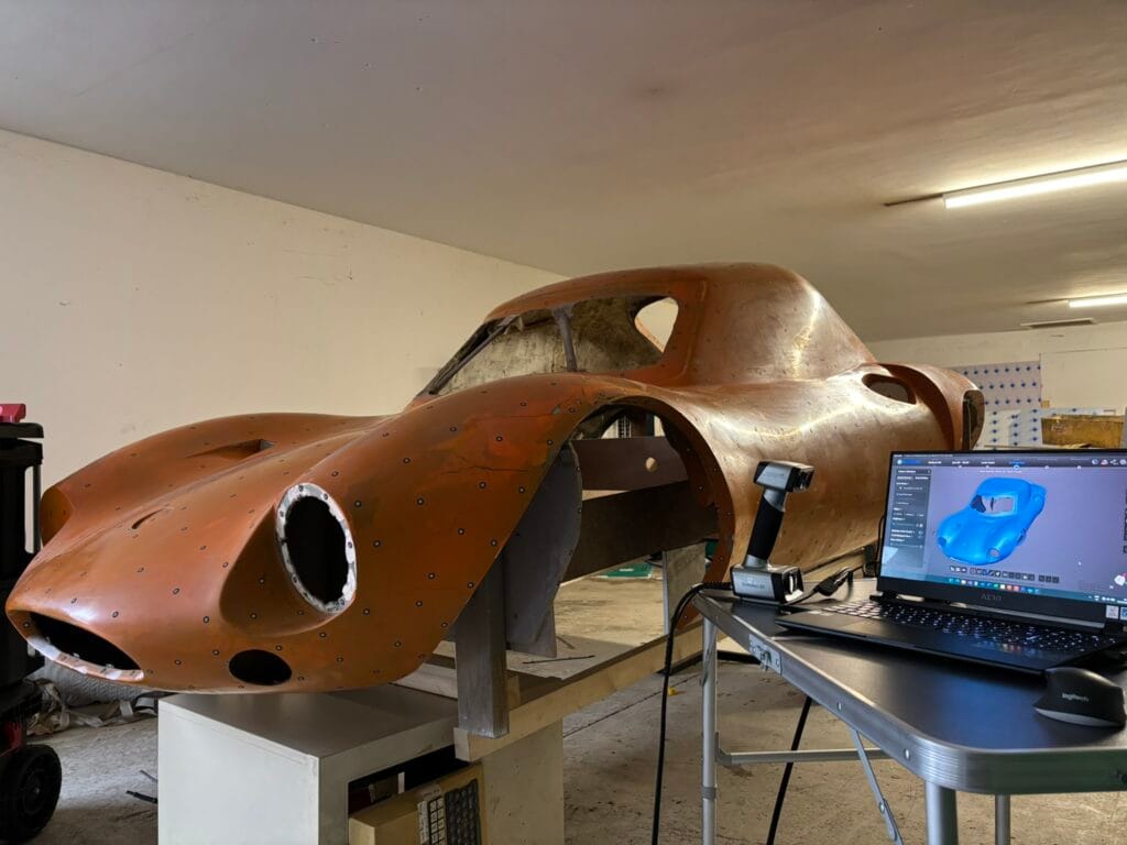Orange fibreglass car body on supports beside a laptop and 3D scanner, with the 3D scan visible on the laptop screen.