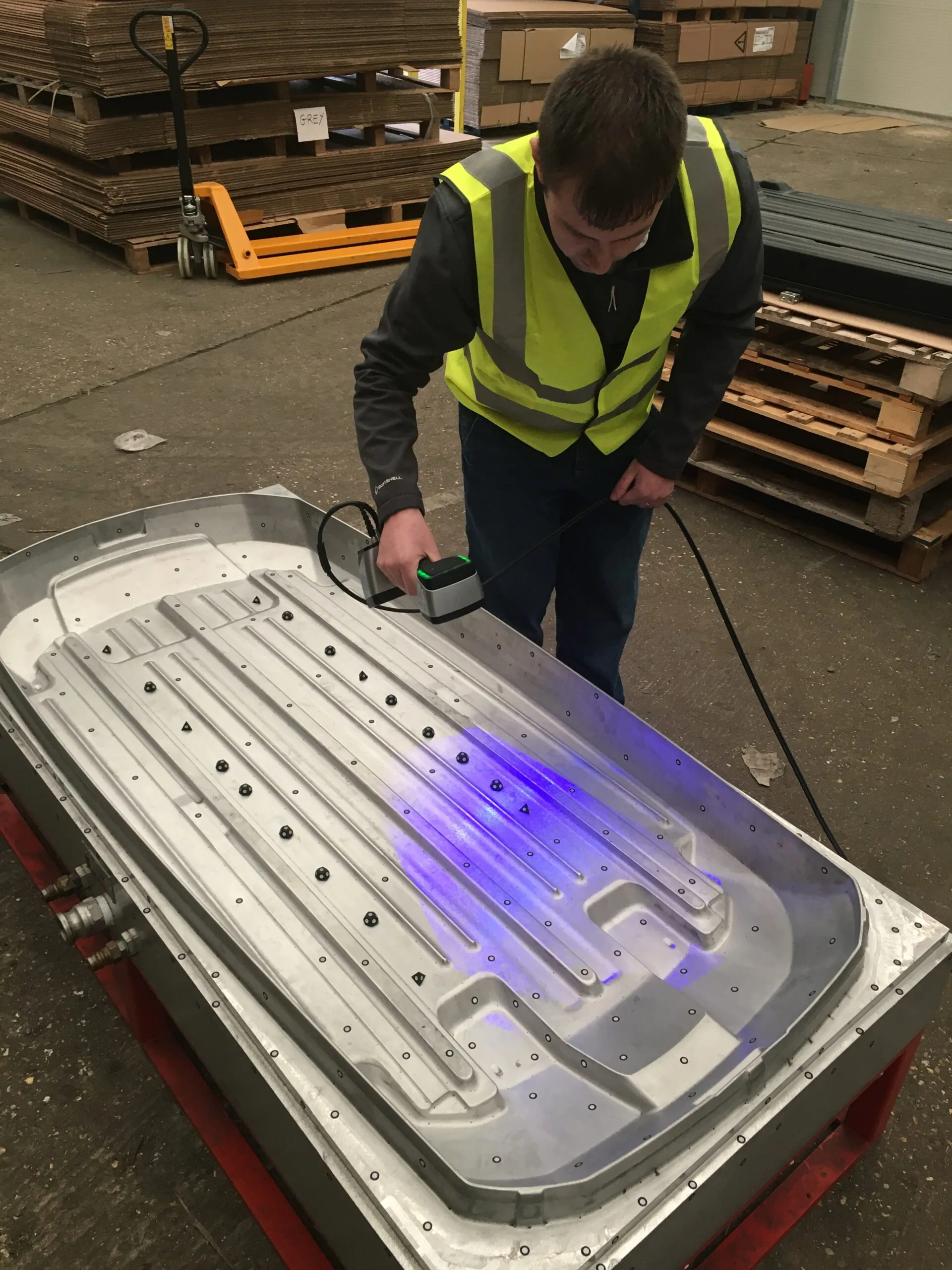 Man in yellow high-visibility clothing scanning a large silver mould tool in a factory warehouse.